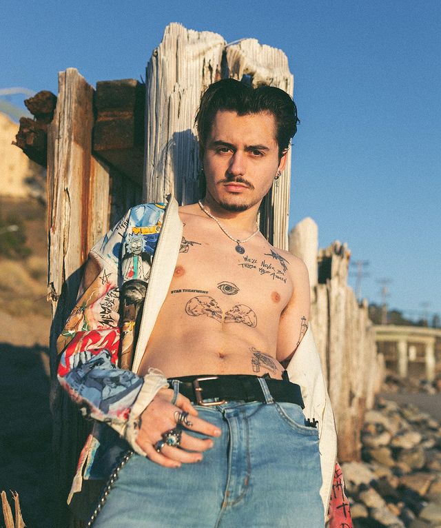 Isaak Presley in a colorful shirt and blue jeans showing his tattoos.